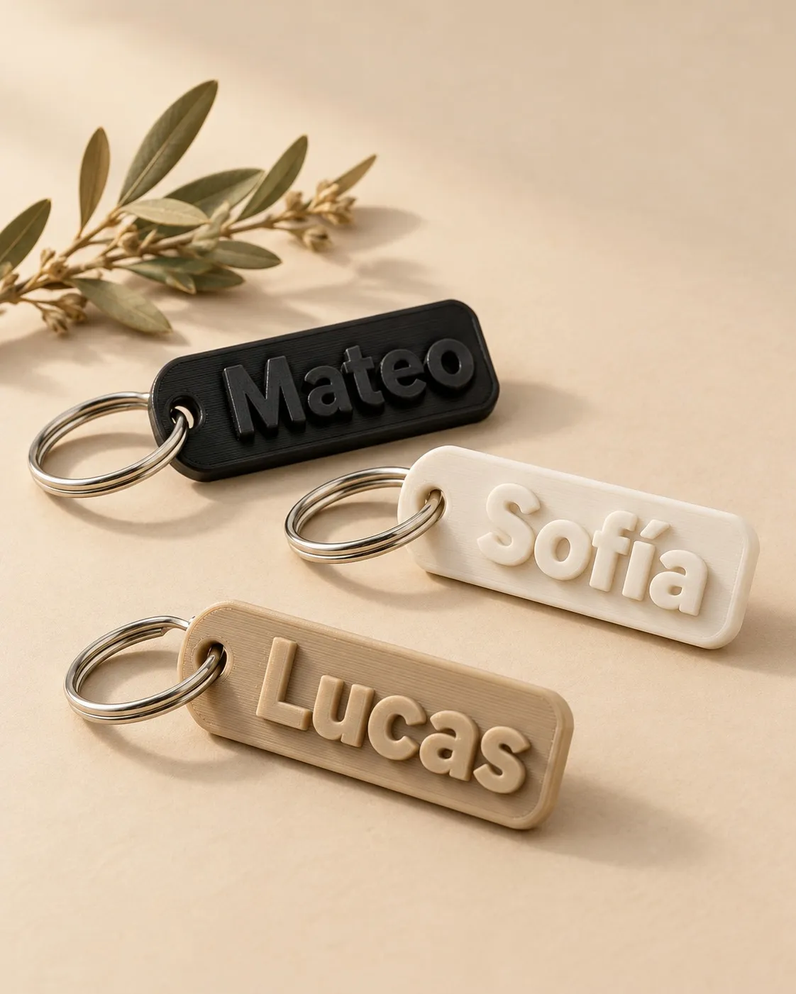 Tres llaveros impresos en 3D con nombres en relieve, en negro, blanco y beige, sobre fondo beige.
